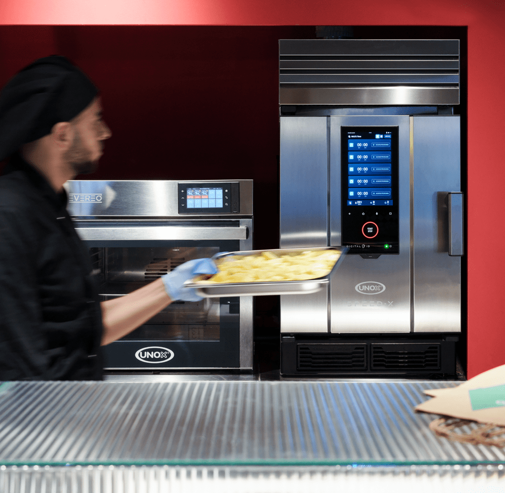 QSR operator walking in front of an unox speedx speed oven and evereo hot fridge with a tray of potatoes in his hand.